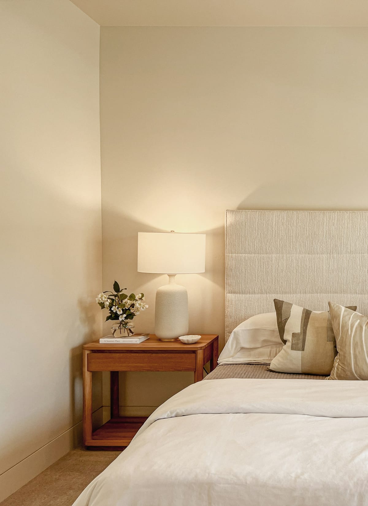 A cream table lamp glowing on a wooden nightstand beside a soft tufted headboard