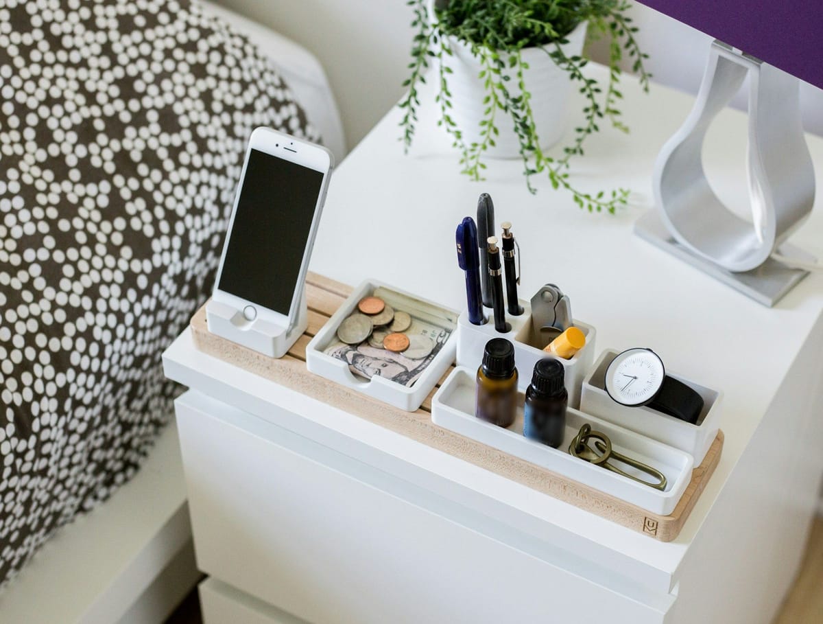 A white nightstand with a wooden organizer tray holding a phone, pens, a watch, and small bottles, beside a trailing plant